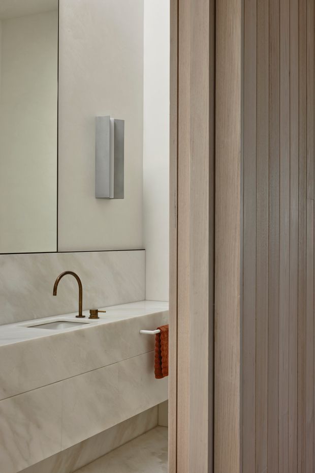 A refined contemporary bathroom viewed through a partially open vertical timber slatted door or screen. The room features a pale stone-clad floating vanity with integrated basin, a large frameless mirror, a minimalist rectangular wall light, and a curved bronze or brass gooseneck tap with matching controls. A small rust-coloured hand towel adds warmth against the neutral stone and light timber palette.