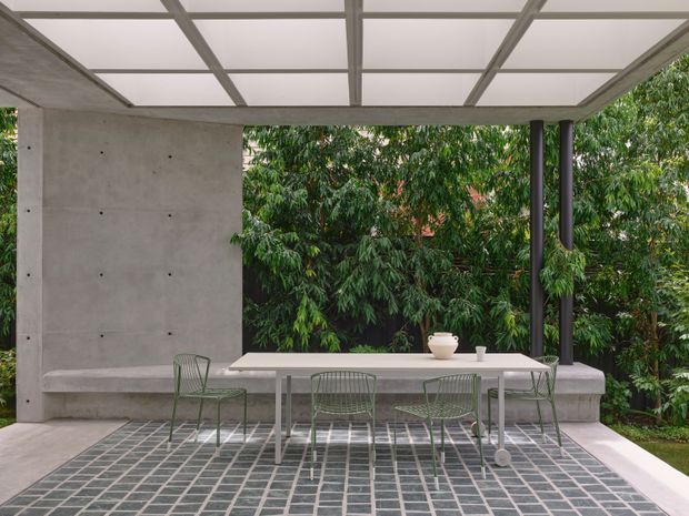 A minimalist covered outdoor dining terrace with a long light-colored dining table and wire-frame chairs, set against a concrete wall and dense greenery. The patio features a translucent grid roof, slender dark posts, and a geometric grid-patterned tiled floor, creating a refined indoor-outdoor entertaining space.