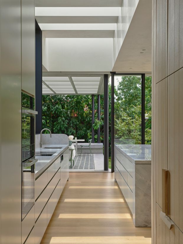 A refined contemporary kitchen viewed along a narrow galley layout toward a glazed outdoor entertaining area. The kitchen features sleek light cabinetry, integrated appliances, a pale stone island or benchtop, timber flooring, dark-framed floor-to-ceiling glazing, and a timber-lined wall. Exposed ceiling beams and a covered terrace with outdoor seating are visible beyond.