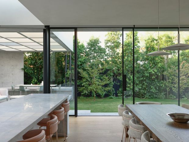 A refined contemporary open-plan kitchen and dining area opening onto a landscaped garden through extensive floor-to-ceiling glazing. The kitchen features a large pale stone island with curved stools, while a long timber dining table with upholstered chairs occupies the foreground. Black-framed sliding or folding glass doors, a covered outdoor area, pendant lights, and a calm neutral palette create a bright indoor-outdoor connection.