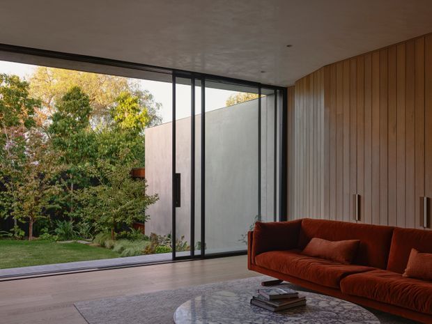 A refined contemporary living room with a low-profile rust-red sofa, marble coffee table, and large-format rug. Full-height black-framed sliding glass doors open the room to a landscaped garden and concrete courtyard wall, while the interior features warm vertical timber wall panelling, a smooth light ceiling, and pale timber flooring. The room has a strong indoor-outdoor connection and a calm, warm, sophisticated atmosphere.