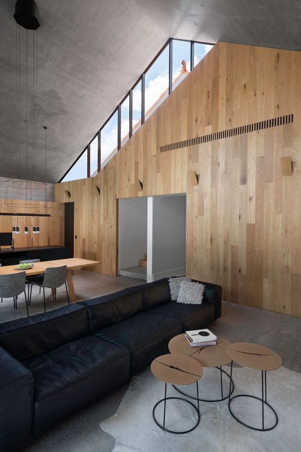 A spacious open-plan interior with a living room in the foreground, featuring a large black leather sectional sofa and nested round coffee tables. Behind it is a timber-lined dining and kitchen area with a dining table, upholstered chairs, dark cabinetry, and pendant lighting. The room has a dramatic sloping concrete ceiling, tall triangular clerestory glazing, exposed timber wall lining, polished concrete flooring, and a stair opening or passage in the rear wall. The overall design is warm, refined, and contemporary with strong natural-material contrast.