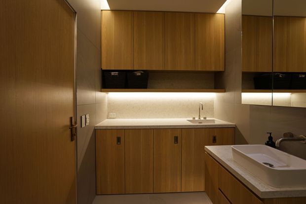 A compact, warmly lit bathroom with extensive timber cabinetry and wall panelling, a pale stone benchtop and splashback, integrated under-cabinet lighting, a vanity basin in the foreground, and a mirrored cabinet reflecting the room. A timber door is visible on the left.