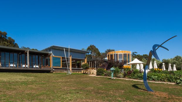 Wide exterior view of a low-rise contemporary property set in landscaped grounds. The building combines dark horizontal cladding, extensive floor-to-ceiling glazing, timber-framed elements, and a prominent curved cylindrical glazed volume. A sculptural metal artwork, garden planting, umbrellas, and a white outdoor canopy occupy the lawn and terrace area under a clear blue sky.