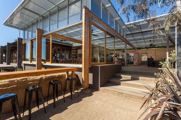 Exterior view of a contemporary hospitality-style outdoor dining and entertaining area, featuring extensive floor-to-ceiling glazing, exposed timber posts and beams, translucent roof canopies, built-in bar seating, steps, and a covered courtyard-like space. The warm timber and earthy finishes contrast with dark metal stools and transparent glass.
