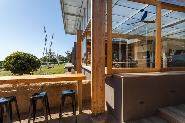 A bright semi-enclosed outdoor dining or entertaining area beside a glazed building facade. Warm timber posts and window frames support a transparent roof and large windows, with a built-in timber bar ledge, dark stools, a rendered masonry wall, and a visible interior with a ceiling fan. The area opens onto landscaped grounds and receives abundant natural light.