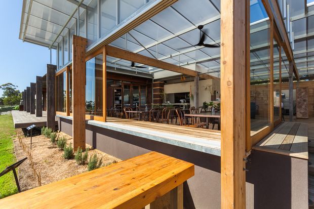 A contemporary timber-framed building with extensive floor-to-ceiling glazing, a translucent roof canopy, and a spacious indoor-outdoor dining and entertaining area. A large timber table occupies the foreground, while timber columns, exposed beams, outdoor seating, and a visible kitchen/bar area create a warm, connected pavilion-like space.