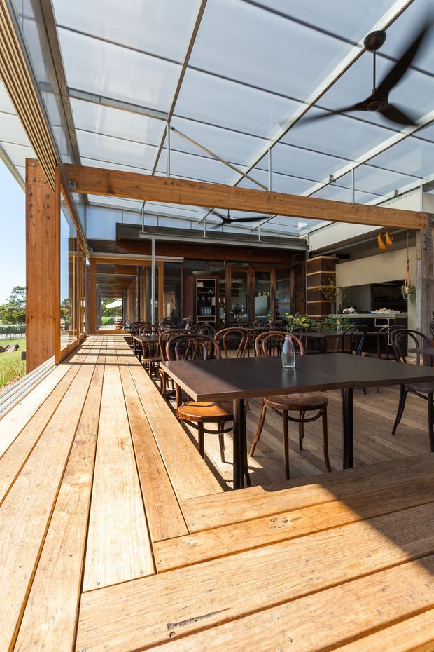 A spacious semi-outdoor dining area with a long dark dining table and bentwood chairs, enclosed by extensive timber decking and framed with exposed timber beams and posts. The space has a translucent glazed or polycarbonate roof, large open glazing, a ceiling fan, and a built-in bar or kitchen area at the rear. It opens onto landscaped grounds and has a warm, naturally lit hospitality-style atmosphere.