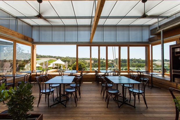 A bright, glazed dining or hospitality space with exposed timber beams, timber flooring, expansive windows, and multiple dark metal café tables and chairs. The room opens visually to a landscaped outdoor setting and appears designed for casual dining, with built-in shelving or service joinery along the right wall.