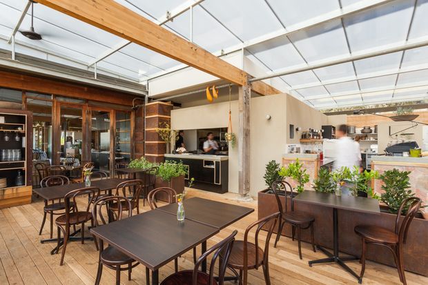 A bright, open-plan café or restaurant interior with dining tables and bentwood chairs in the foreground, a visible service counter and commercial kitchen, extensive glazing, timber ceiling beams, and a translucent roof structure. The space combines warm timber finishes with dark metal and earthy cabinetry, creating an industrial-contemporary hospitality setting.