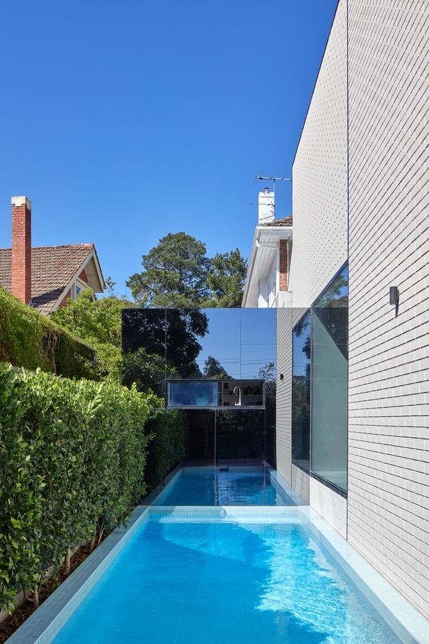 Exterior view of a narrow urban residential property featuring a long blue swimming pool confined between a dense hedge and a tall contemporary white brick-clad wall. Large reflective glazing and dark glass panels connect the pool to the house, while neighbouring traditional homes with tiled roofs and brick chimneys are visible beyond the boundary. Bright clear daylight and strong geometric lines dominate the composition.