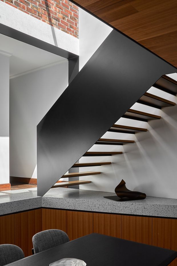 A refined interior view centred on a sculptural open-riser staircase with a dramatic matte-black structural side panel. The staircase rises beside white plaster walls and an exposed red-brick section, beneath a warm timber ceiling. A speckled stone benchtop and dark timber cabinetry form a built-in counter or dining area in the foreground, with black dining furniture and a small sculptural object. The composition combines strong geometric lines, contrasting black and white surfaces, warm wood, and heritage architectural elements.