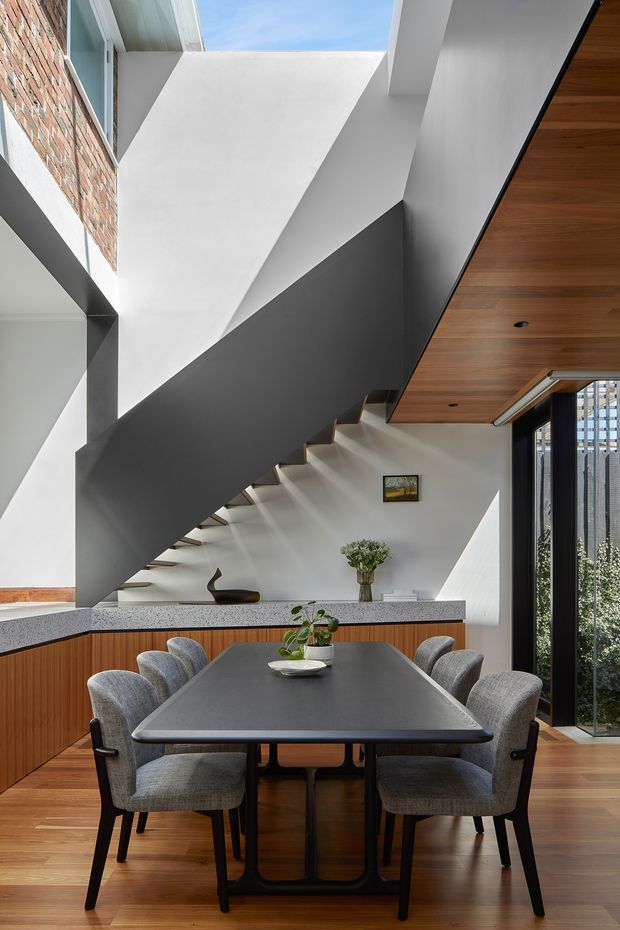 A refined contemporary dining area beneath a dramatic sculptural staircase. The room features a large dark dining table with grey upholstered chairs, warm timber flooring and a timber-lined ceiling, built-in timber cabinetry, white plaster walls, a dark charcoal stair soffit, exposed brickwork, and tall glazed doors or windows opening to greenery. Strong natural light enters from above and from the glazed side.