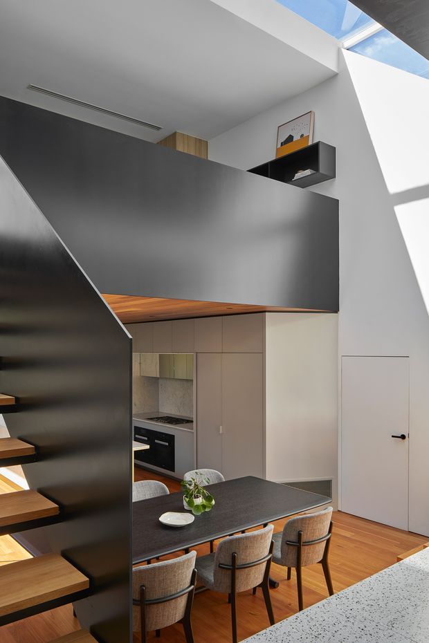 A vertically composed view of a compact contemporary kitchen and dining area beneath a dramatic open-riser staircase. Matte charcoal stair and upper partition walls contrast with warm timber stair treads and flooring, pale integrated cabinetry, and a dark dining table with upholstered chairs. A skylight and high-level glazing bring strong natural light into the double-height space. The kitchen includes built-in appliances, a timber benchtop, and a stone-look foreground counter.
