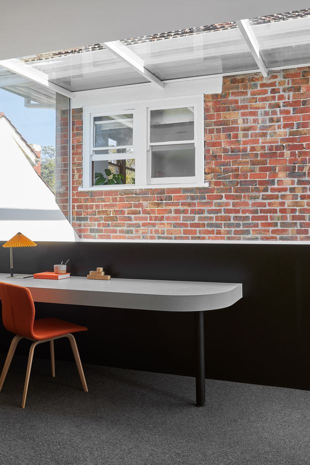 A compact, contemporary home office with a long curved-edge white desk set against a matte black wall. An orange upholstered chair, small orange desk lamp, books and decorative objects add colour. Above the desk, a white-framed window looks onto an exposed red brick exterior wall beneath a translucent covered roof structure. Dark carpet and strong natural light create a crisp, graphic interior.