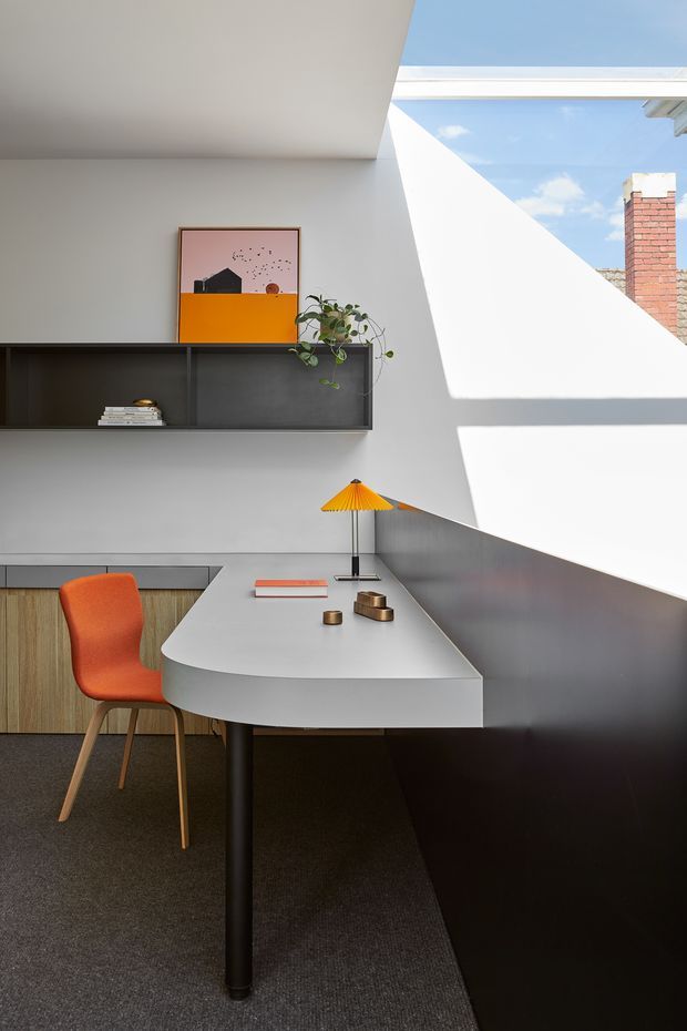 A bright, compact home office nook with a curved white built-in desk, black shelving and cabinetry, timber drawer fronts, and a vivid orange upholstered chair. A small yellow desk lamp, books, artwork, and a trailing plant add colour. Strong sunlight enters through a large angled skylight or clerestory opening, creating geometric shadows across the white walls and dark cabinetry.