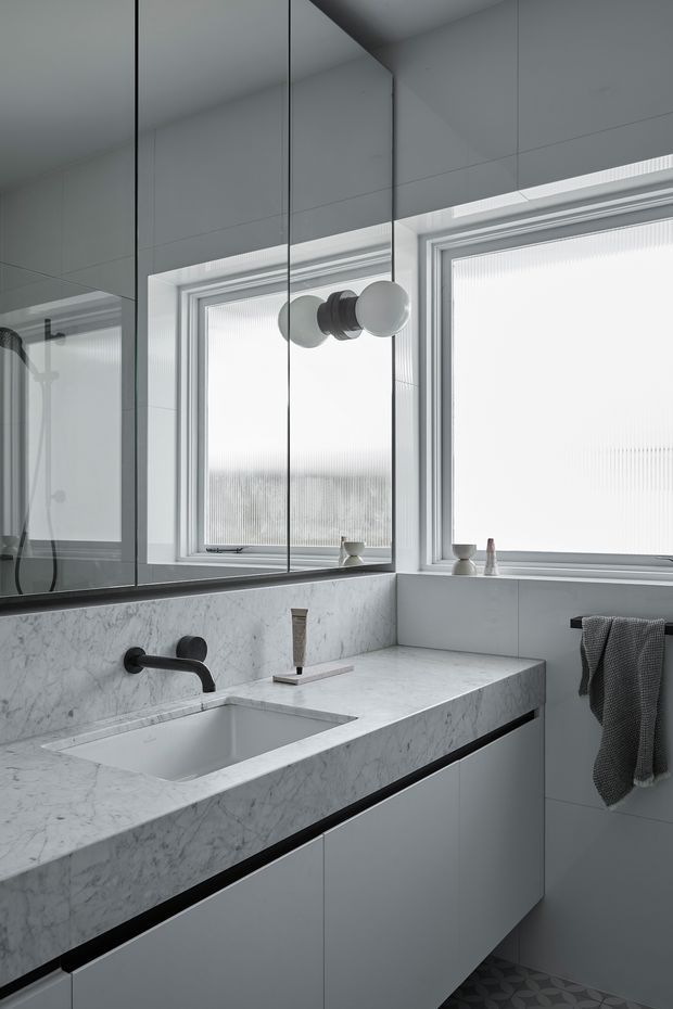 A refined contemporary bathroom vanity in a compact, bright space. The room features a floating pale grey vanity with an integrated rectangular basin, a white marble-look benchtop and splashback, matte black wall-mounted tapware, large mirrored cabinetry or wall panels, frosted ribbed windows, and dark patterned floor tiles. The restrained monochromatic palette creates a clean, calm atmosphere.