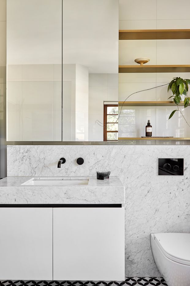 A compact contemporary bathroom with a wall-mounted white vanity, integrated rectangular basin, black wall-mounted tapware, and a freestanding white toilet. White marble-look stone cladding covers the vanity benchtop and wall, while glossy white cabinetry and large white wall panels create a clean, minimalist backdrop. A patterned black-and-white floor adds contrast. Timber-framed glazing and open timber shelves with restrained styling are visible in the upper portion of the room.