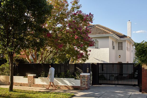 Street-facing view of a refined two-storey white rendered home behind a black vertical-slat security fence and sliding driveway gate. The house has a hipped terracotta-tiled roof, white-framed windows with louvred/shutter-like detailing, and a prominent white chimney. Brick gate piers, a paved driveway, mature flowering trees, and landscaped frontage complete the scene.