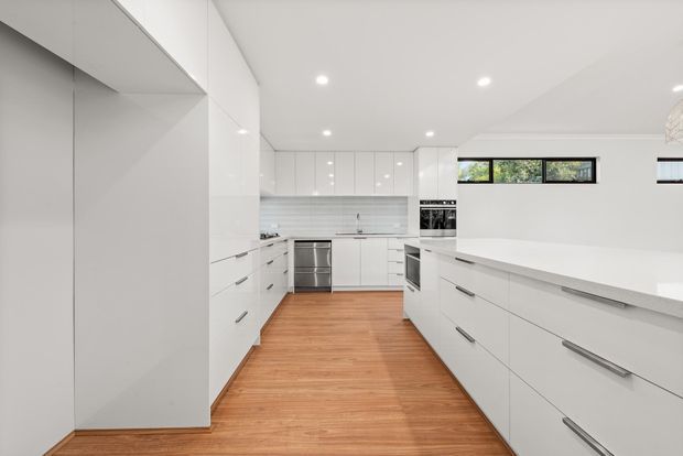 Bright, spacious contemporary white kitchen with extensive handle-fitted cabinetry, a large island or peninsula with a white benchtop, integrated appliances, and warm timber flooring. Narrow horizontal windows provide natural light, while recessed ceiling downlights create an even, clean-lit interior.