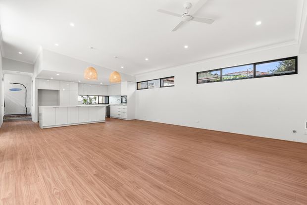 Bright, open-plan contemporary interior featuring a white kitchen with a long island, integrated cabinetry, warm timber-look flooring, high horizontal windows, recessed ceiling lights, a white ceiling fan, and two woven pendant lights. The largely unfurnished area appears suitable as a combined living and dining space adjoining the kitchen.