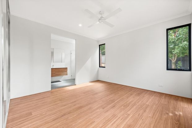 Bright, minimally furnished bedroom with warm timber flooring, white walls and ceiling, black-framed windows, recessed downlights, and a white ceiling fan. An open doorway reveals part of an adjoining bathroom with a timber vanity and grey floor.
