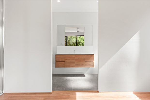 Bright minimalist bathroom viewed through a wide opening, featuring a floating timber vanity with integrated white basin, large rectangular mirror, grey tiled floor, white walls, and strong natural sunlight. Timber flooring in the foreground and a reflected window view add warmth and contrast.