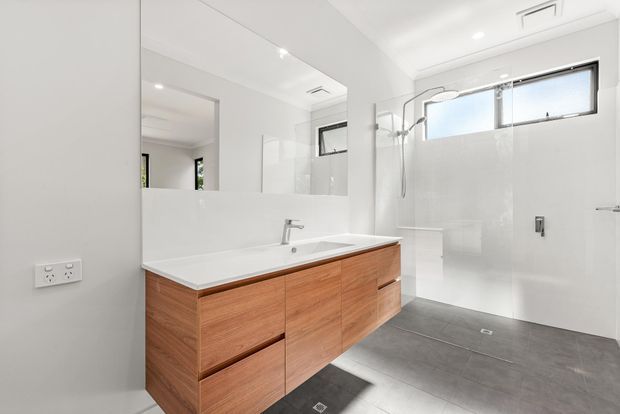 Bright contemporary bathroom with a large timber vanity, white countertop and integrated basin, expansive wall mirror, frameless glass shower enclosure, white tiled walls, grey floor tiles, and high horizontal windows. The clean-lined design is spacious and minimal with warm timber cabinetry providing contrast.
