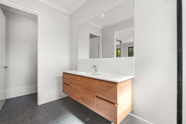 Bright, contemporary bathroom featuring a floating timber-veneer vanity with white benchtop and integrated basin, a large wall-to-wall mirror, light grey walls, white trim, and dark grey floor tiles. An open doorway is visible to the left.