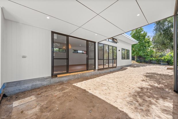 A bright modern covered outdoor area or courtyard beside a single-storey extension. The building has white vertical wall cladding, dark-framed floor-to-ceiling glazed sliding doors and windows, a white panelled ceiling with recessed downlights, and a concrete plinth. The sandy ground and surrounding trees are visible beyond the glazed facade.
