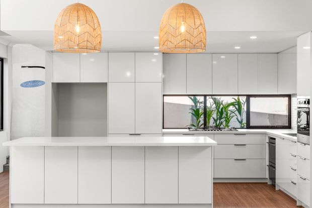 Bright contemporary white kitchen with extensive handleless cabinetry, a large island with a white benchtop, integrated appliances, timber flooring, and two prominent woven pendant lights. A horizontal window with indoor plants provides a natural accent.