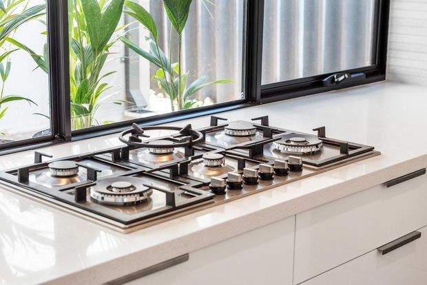 Bright close-up of a modern kitchen cooktop with five gas burners set into a light engineered-stone countertop. White handleless cabinetry with dark linear pulls sits below, while black-framed windows provide abundant natural light and views of indoor greenery.