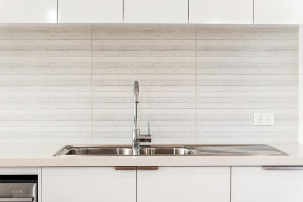 Close-up view of a minimalist kitchen work area with white flat-panel cabinetry, a long stainless-steel sink and drainer, pale stone-look benchtop, and horizontally textured light tile splashback. A chrome pull-out mixer tap and power outlet are visible.