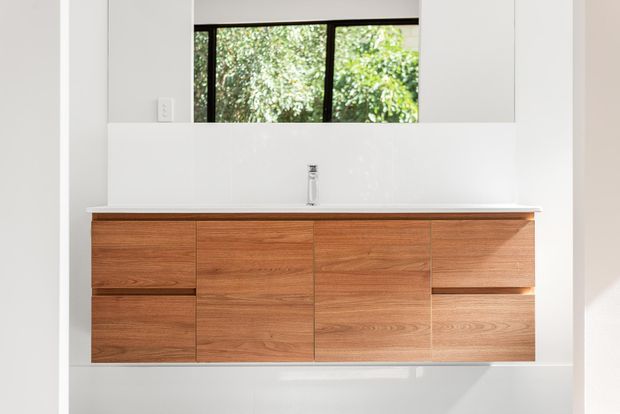 Minimalist bathroom vanity with a wall-mounted timber cabinet featuring multiple drawers and doors, a white benchtop with integrated basin and chrome tapware, and a large mirror above. A horizontal black-framed window reveals greenery outside, while white walls and surfaces create a bright, clean composition.