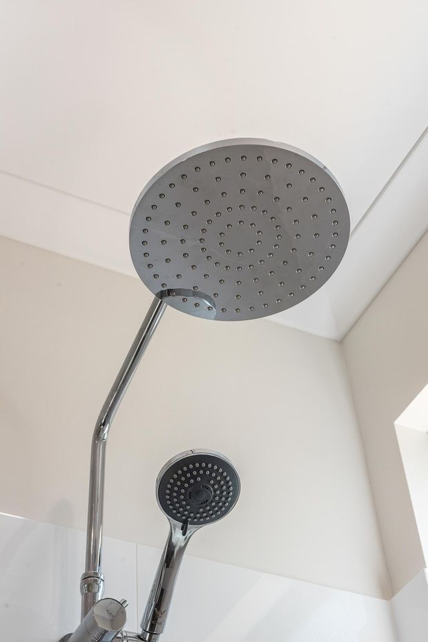 Close-up view of a modern chrome shower system in a bathroom, featuring a large round overhead rain shower and a handheld shower on a vertical rail. The shower is set against clean white wall and ceiling surfaces with minimal visible detailing.