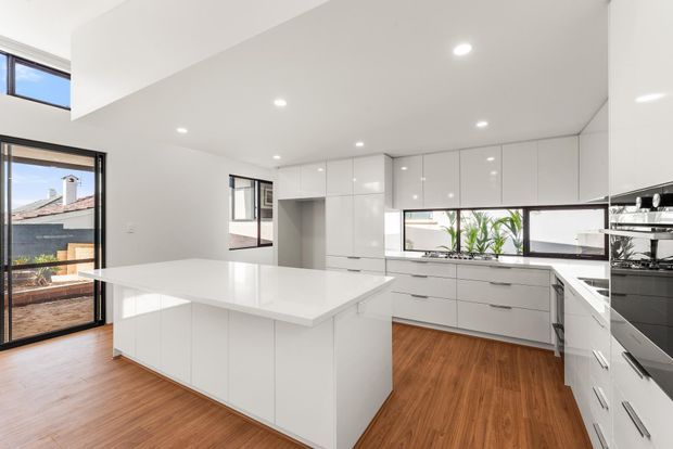 Bright contemporary white kitchen with extensive glossy built-in cabinetry, a large central island, white benchtops, integrated appliances, and warm timber flooring. Large windows and glazed doors provide abundant natural light and views to neighbouring rooftops and greenery.