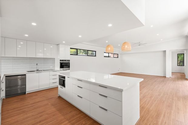 Bright open-plan kitchen and living area with a large white island, extensive white flat-panel cabinetry, integrated appliances, tiled splashback, warm timber flooring, and woven pendant lights. The space has a clean contemporary feel with abundant recessed lighting and high ceilings.