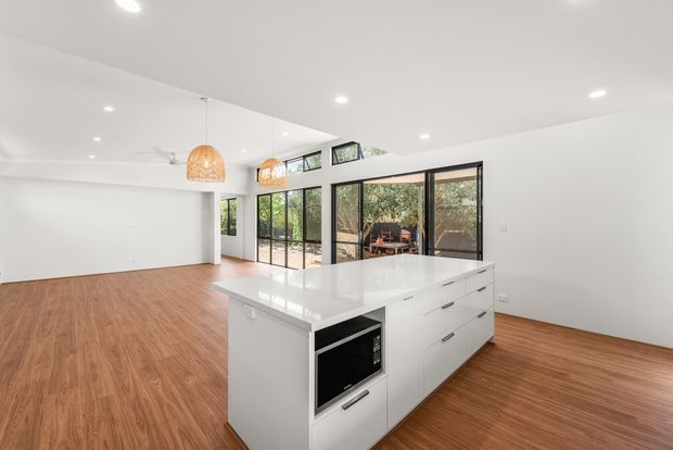 Bright open-plan contemporary kitchen with a large white island, integrated appliances and extensive black-framed glazing. Warm timber flooring, recessed ceiling lighting and woven pendant lights create a clean but inviting interior, with a dining or outdoor area visible beyond the glazing.
