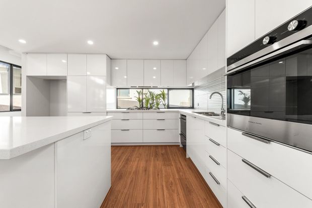 A bright, contemporary white kitchen with extensive glossy full-height cabinetry, a large white island, integrated double ovens, stainless-steel sink and tap, and a warm timber-look floor. Horizontal windows provide natural light above the rear benchtop, while recessed ceiling lights and clean-lined hardware reinforce the streamlined design.