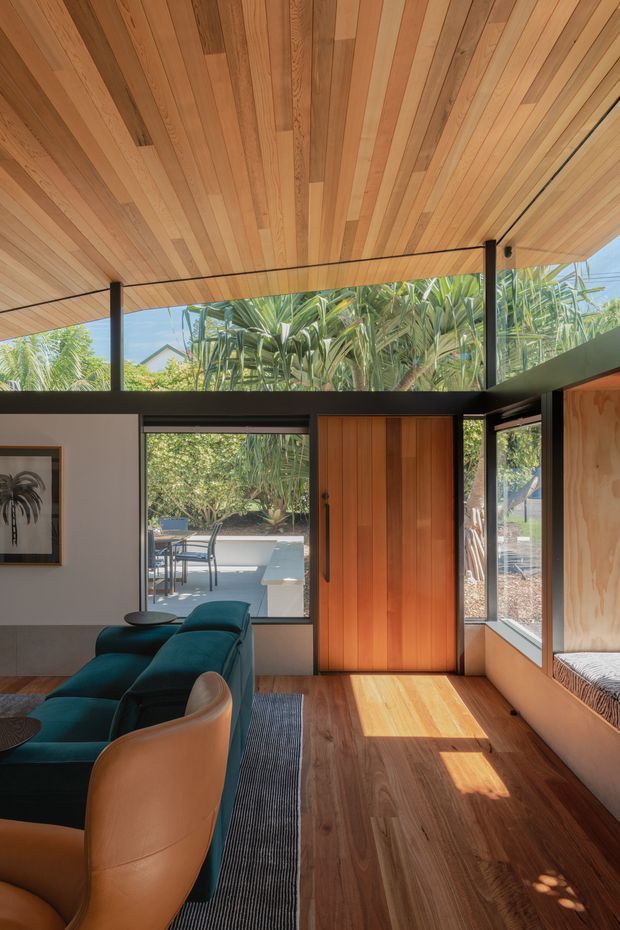 A warm, light-filled contemporary living room with extensive glazing opening toward a leafy outdoor patio. The room features a timber-lined sloping ceiling, dark slender structural framing, timber flooring, a rich timber entry door, built-in window seating, and a deep teal sectional sofa with a tan leather lounge chair. Sunlight creates strong patterns across the floor, while tropical planting is visible outside.