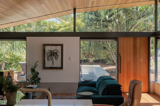 A bright, tropical living room with expansive floor-to-ceiling glazing overlooking lush planting and a small outdoor pool or water feature. The room features a warm timber-lined ceiling, dark window frames, a white plaster feature wall with framed botanical artwork, a deep teal sofa, a timber entry door, and contemporary furnishings. The open connection to the garden creates an airy indoor-outdoor atmosphere.