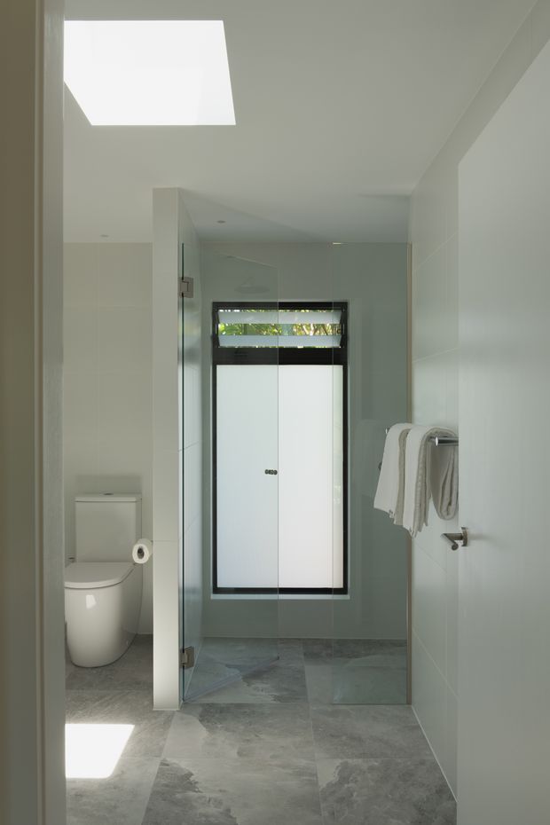 Bright modern bathroom with pale tiled walls, large grey stone-look floor tiles, a glass-enclosed shower, wall-mounted towel rail, toilet, skylight, and a narrow black-framed window providing filtered daylight.