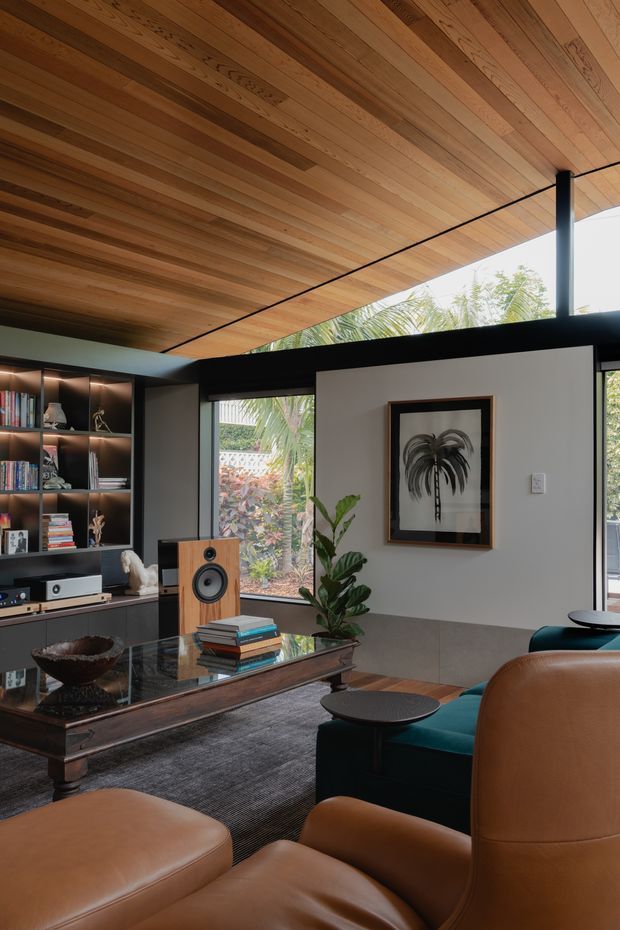 A warm, sophisticated living room with a strong indoor-outdoor connection. The room features a richly timber-lined ceiling, black-framed glazing, dark built-in shelving and media joinery, a wood-toned speaker, a glass-topped coffee table, leather lounge chairs, and a deep teal sofa. Tropical planting is visible through the windows, while framed botanical artwork and layered furnishings add character.