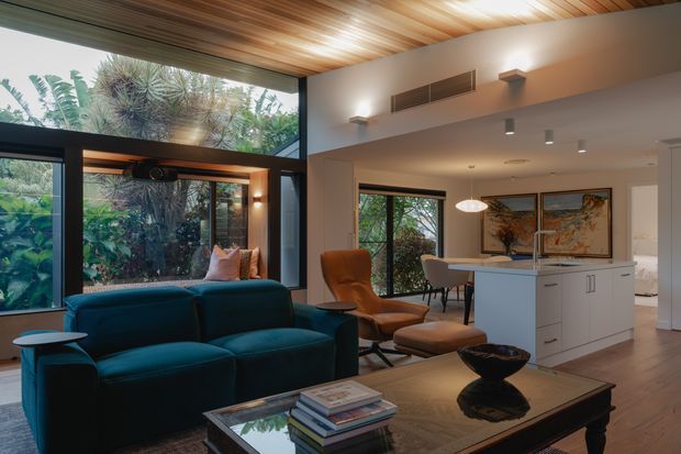 A warm, refined open-plan living area with a deep teal sofa, leather lounge chair and ottoman, and a dark coffee table with books and decor. Floor-to-ceiling glazing frames lush garden views, while a timber-lined ceiling adds warmth. A white kitchen island and dining area are visible in the background, with contemporary artwork and restrained lighting throughout.