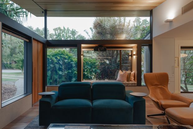 A refined living room with floor-to-ceiling glazing opening onto lush greenery. The room features a deep teal sofa, cognac leather lounge chair and ottoman, dark timber-framed windows, a warm timber ceiling, built-in window seating, and integrated wall lighting. The high glazing and layered timber detailing create a calm, nature-connected contemporary interior.