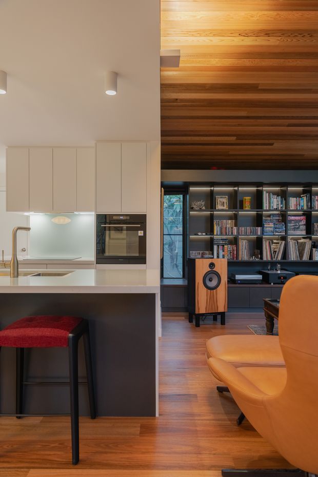 A refined open-plan interior combining a contemporary kitchen and living area. The kitchen features pale flat-panel cabinetry, integrated appliances, a light benchtop, and a dark island with a red upholstered bar stool. The adjoining living space has warm timber flooring and a dramatic timber-lined ceiling, with full-height dark built-in shelving filled with books and media equipment. A freestanding timber speaker and mustard-orange leather lounge chair add strong mid-century character.