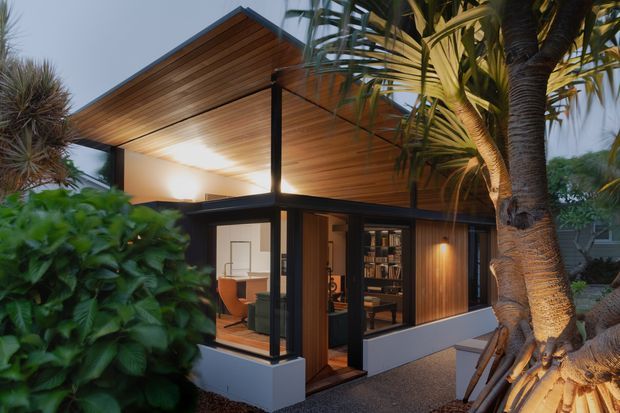 A compact modern pavilion-style dwelling set within lush tropical landscaping. The building features a dramatic asymmetrical pitched roof with a warm timber-lined soffit, dark slender structural framing, extensive floor-to-ceiling glazing, timber wall cladding, and a raised pale base. Interior furnishings and shelving are visible through the windows and open entry, while palms and dense planting frame the exterior.