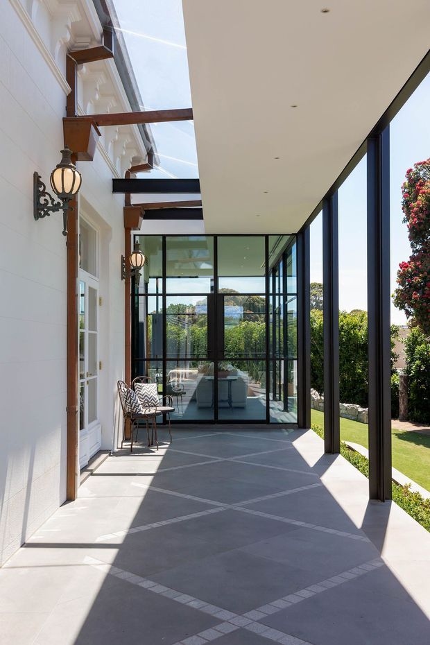A bright covered outdoor terrace runs alongside a white traditional-style house, combining ornate wall lanterns and timber beam details with a sleek black-framed glazed extension. The terrace has a large geometric tiled or paved floor pattern, while the glass enclosure reveals a furnished interior beyond. Manicured gardens and clipped shrubs are visible outside, with strong sunlight and deep shadows creating a high-contrast, refined setting.