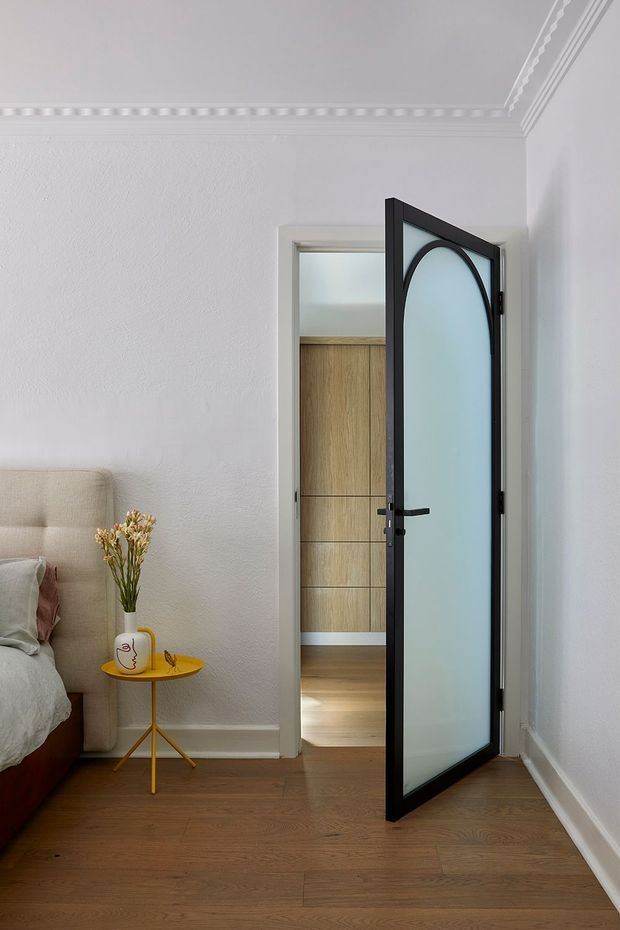 A refined bedroom interior with warm timber flooring, white textured walls, decorative ceiling cornice, and a partially visible upholstered bed. An open black-framed door with a tall arched frosted-glass panel leads to a timber-lined adjoining space, likely a wardrobe or dressing area. A small mustard-yellow side table with a vase adds a subtle accent.