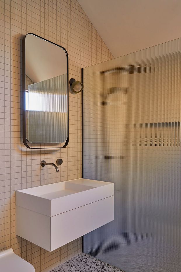 A compact contemporary bathroom with a wall-mounted white floating vanity and integrated basin, brushed bronze wall-mounted tapware, a rounded-rectangle black-framed mirror, small beige grid tiles covering the walls, and a vertically ribbed translucent glass shower partition. The floor appears to be terrazzo-like with dark aggregate. Warm ambient lighting creates a refined, softly muted atmosphere.